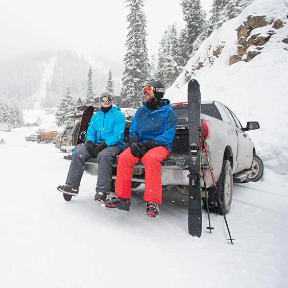 two skiers in the snow sitting on the tailgate of a truck