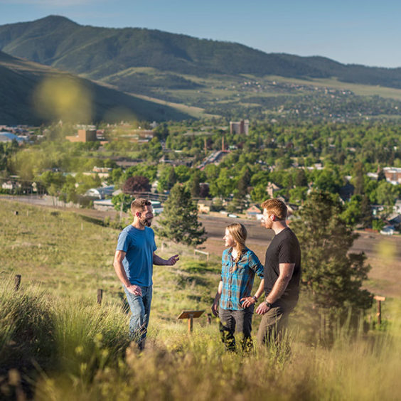 Two men and a woman standing on a hill talking with the Missoula valley in the background.