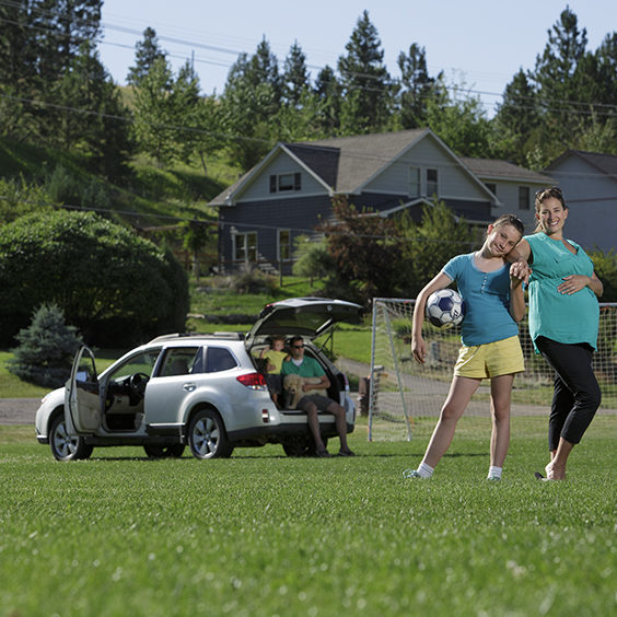 Girl holding soccer ball, standing next to pregnant mom with car in the background.