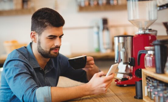 Man sitting a counter using a cell phone while drinking a coffee.