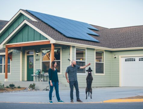 Man and woman walking in front of their house while their dog is doing a trick.