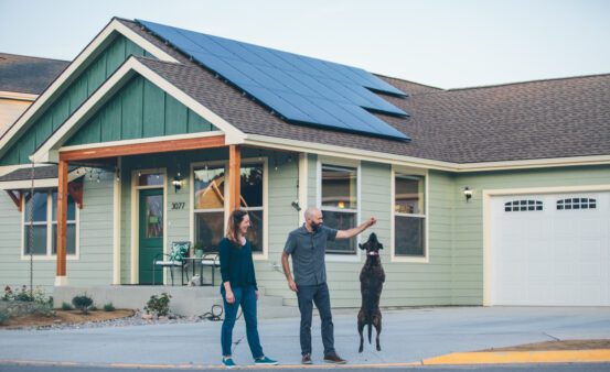 Man and woman walking in front of their house while their dog is doing a trick.