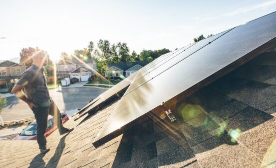 Man standing on the roof of his house next to solar panels during sunset.