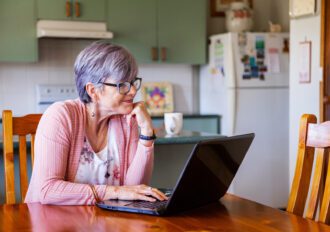 Older woman looking at her laptop while sitting in her kitchen at a table.