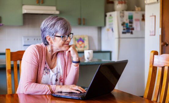 Older woman looking at her laptop while sitting in her kitchen at a table.