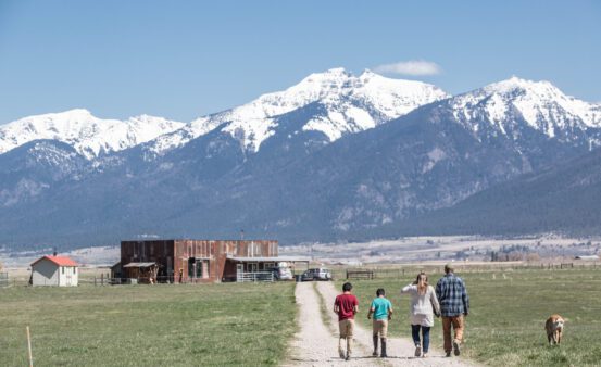 Man and woman walk with their two kids and dog down a country road towards their house with the Mission Mountains in the background.