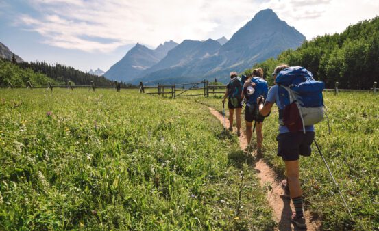 Three hikers with backpacks walking a trail towards the mountains in in summer.