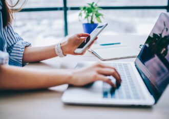 woman's hands holding a cell phone while typing on a laptop sitting at a desk.