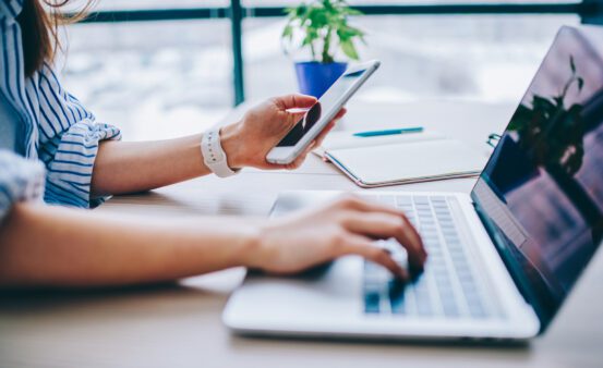 woman's hands holding a cell phone while typing on a laptop sitting at a desk.