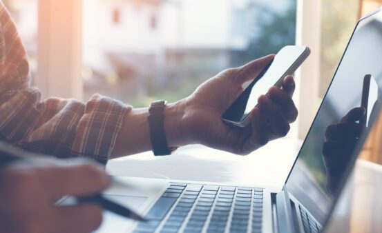 Close up of casual man or freelancer working on laptop computer and holding mobile smart phone with reflection on blank screen, working from home or casual business concept, vintage style.