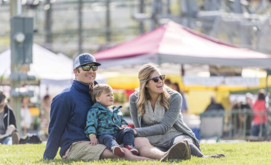 Man and woman sit in the grass with their young child at Missoula out to Lunch at Caras Park in spring.