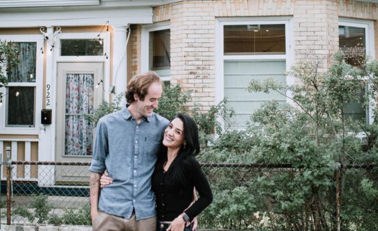 Young man and woman standing on the sidewalk in front of their first house in Butte, Montana.