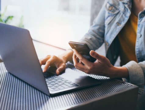 man typing on a laptop computer while holding a cell phone.