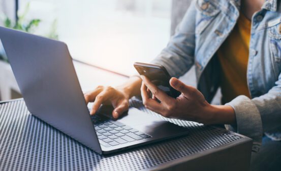 man typing on a laptop computer while holding a cell phone.