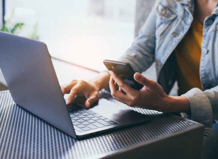 man typing on a laptop computer while holding a cell phone.