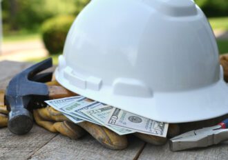 Hard hat sitting on a top of cash, work gloves, and a hammer.