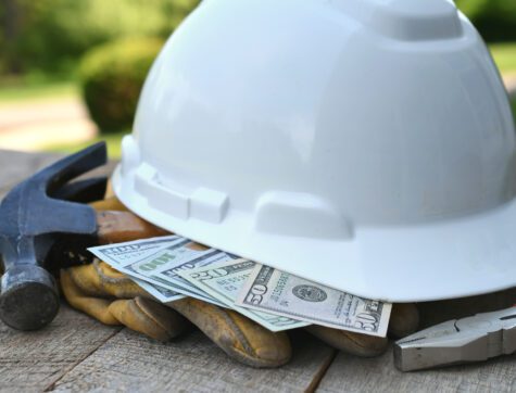 Hard hat sitting on a top of cash, work gloves, and a hammer.