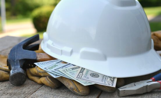 Hard hat sitting on a top of cash, work gloves, and a hammer.