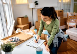 A woman standing at a counter writing on a checklist while surrounded by moving boxes.