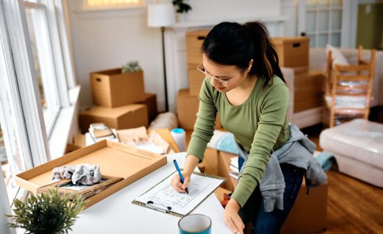 A woman standing at a counter writing on a checklist while surrounded by moving boxes.