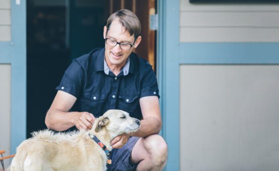 Middle-aged man crouching down to pet his tan dog in front of his house.