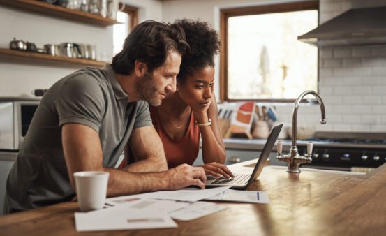 A couple using a laptop to do and financial planning in a kitchen with documents on the counter.