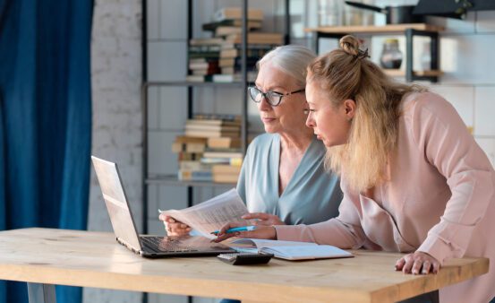 Senior woman using laptop for websurfing in her kitchen. middle-aged daughter helps her mother with documents. Mature lady sitting at work typing a notebook computer in an home office