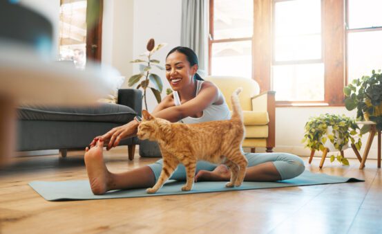 Happy woman doing yoga in her living room with her cat walking by her.