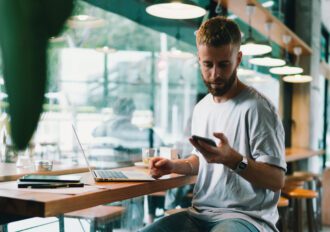 Young trendy male using a mobile phone and a laptop while sitting in a coffee shop.