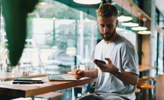 Young trendy male using a mobile phone and a laptop while sitting in a coffee shop.