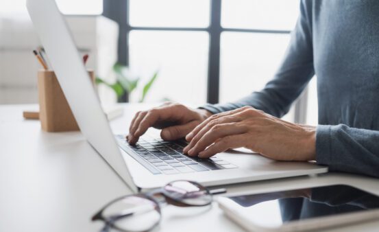 person typing at laptop with phone and glasses near by